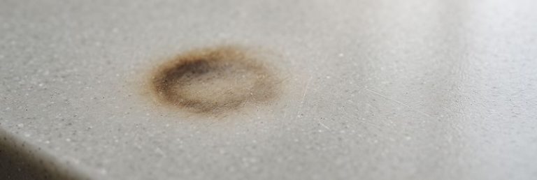 Close-up photo of an acrylic kitchen worktop showing a subtle circular burn mark with darkened edges, surface warping, and faint brown discoloration. Micro-scratches, dust, and debris add texture. The glossy finish reflects soft daylight, while the background remains gently blurred.