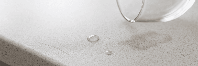 Macro close-up of a Tristone worktop edge showing minor scratches, water droplets, a faint coffee stain, and a tiny glass chip. Natural daylight highlights the fine-grained, matt stone texture, seamless construction, and robust durability against daily wear.