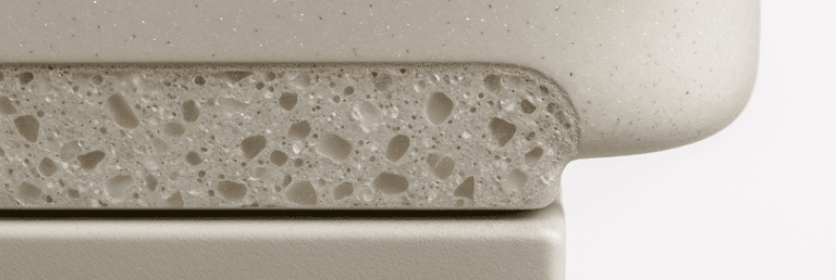 Macro photo of a Hi Macs worktop cross-section showing acrylic resin surface, composite layers, mineral flecks, a polished bullnose edge, and metal support seam, revealing layering, sparkling particles, and craftsmanship against a soft white blurred background.